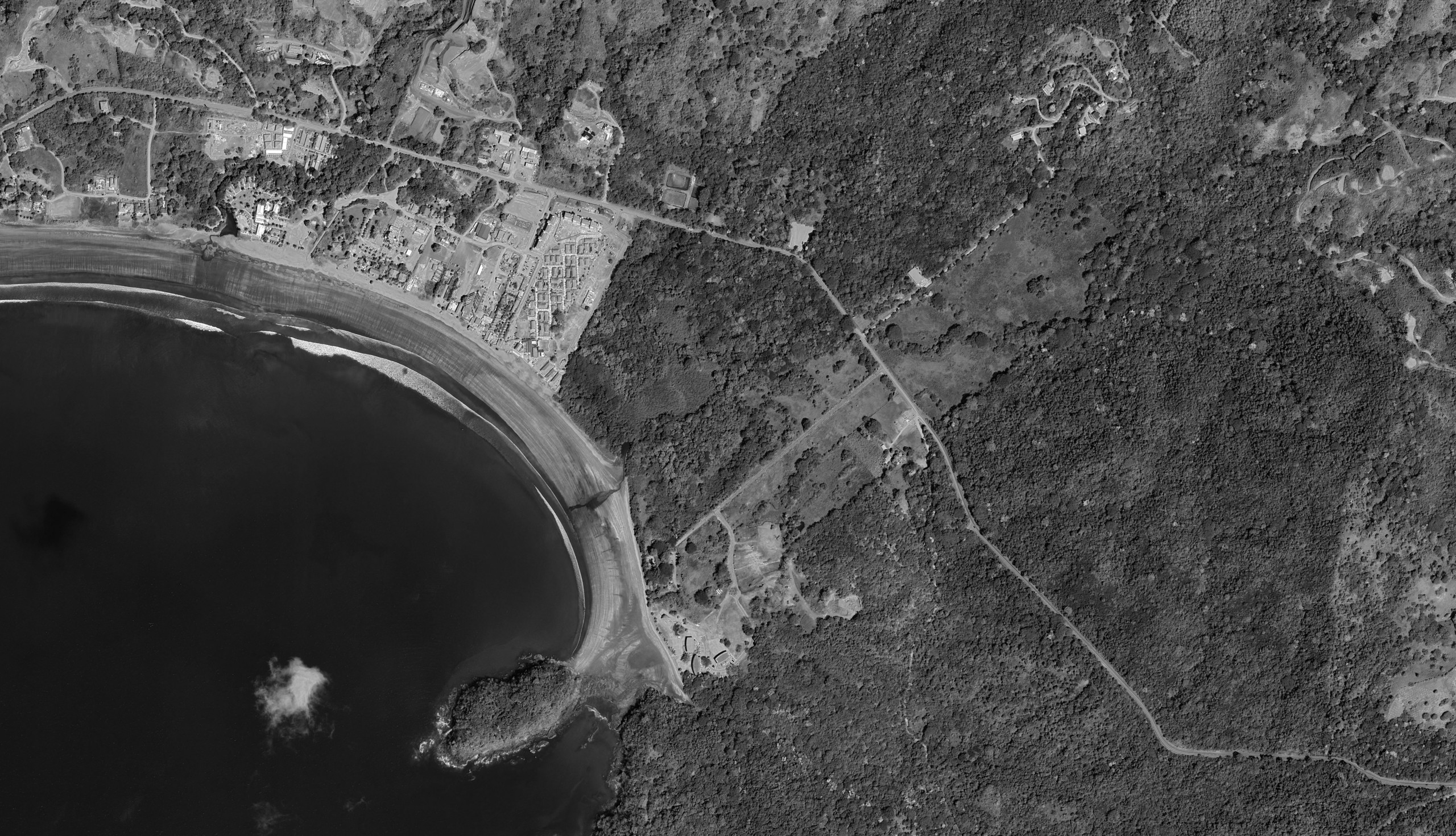 Desaturated aerial photograph of Playa Venao: the dark bay lower left, existing town along the shore, and the forested Cocobolo site extending inland